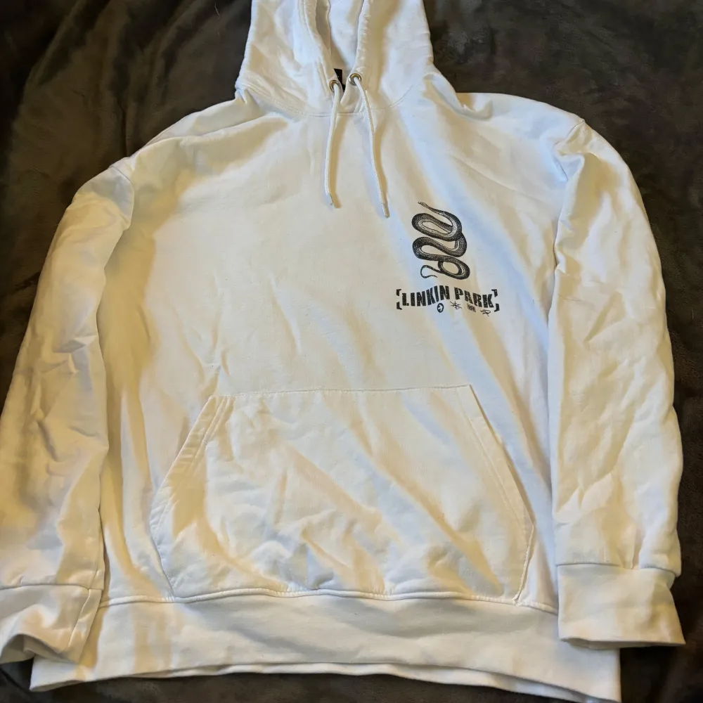 Säljer en vit hoodie med Linkin Park-tryck från Brands In Limited. Den har en stor orm på ryggen och texten 'Linkin Park Los Angeles, California'. Perfekt för fans av bandet! Hoodien har en känguruficka och dragsko i huvan. Skön och stilren design. Köpte den på Zalando.. Tröjor & Koftor.