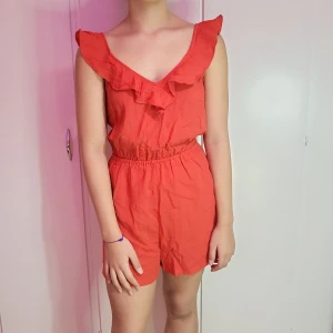 Röd kort jumpsuit med volangdetaljer - Superfin röd jumpsuit med volangdetaljer vid axlarna. Perfekt för sommardagar eller en festlig kväll. Den har en elastisk midja för en bekväm passform. Materialet känns lätt och luftigt, vilket gör den idealisk för varma dagar. Passar perfekt för en avslappnad men stilren look!