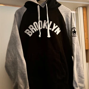 Svart och grå hoodie från Okey Dokey - Säljer en snygg svart och grå hoodie från Okey Dokey med texten 'Brooklyn' på framsidan. Den har en stor ficka fram och justerbara snören i huvan. Perfekt för en avslappnad stil. Materialet är en blandning av bomull och polyester.