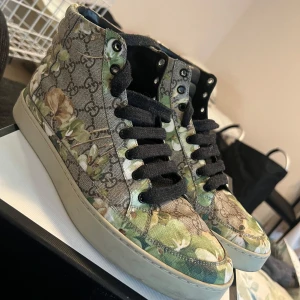 Blommiga sneakers från Gucci - Säljer ett par snygga blommiga sneakers från Gucci i mycket bra skick. Skorna har ett unikt mönster med gröna och beige blommor på en brun bakgrund med Guccis ikoniska logga. Perfekta för att ge din outfit en lyxig touch! Original låda är kvar, dessa skor har bara använts max 10ggr endast till affären dock 😅