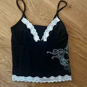 Svart topp med spetsdetaljer - Snygg svart topp med tunna axelband och vackra vita spetsdetaljer längs urringningen och nederkanten. Perfekt för en festlig kväll eller en mysig hemmakväll. Den har en liten rosett framtill som ger en söt touch.