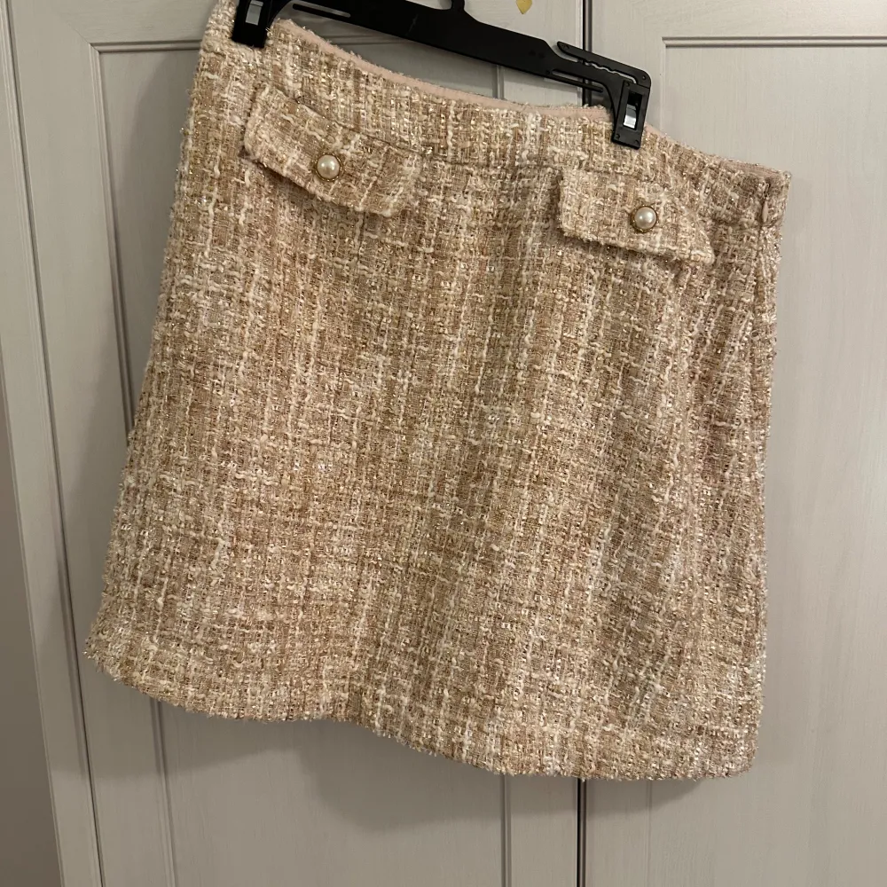 Säljer en superfin beige kjol från Holly & Whyte by Lindex. Kjolen har en elegant textur och dekorativa fickor med pärldetaljer. Perfekt för höst och vår, och kan enkelt kläs upp eller ner beroende på tillfälle. Passar perfekt för en chic och stilren look!. Kjolar.