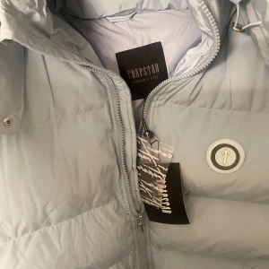 Ljusblå pufferjacka från Trapstar - Säljer en snygg ljusblå pufferjacka från Trapstar. Den har en cool broderad logga på ryggen och en praktisk dragkedja framtill. Perfekt för kalla dagar med sin varma och vadderade design. Jackan har långa ärmar och en stilren look. Passar perfekt för både vardag och chill.