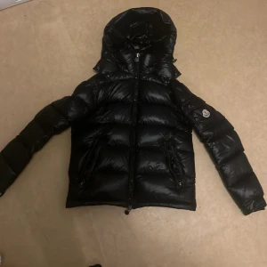 Svart dunjacka från Moncler - Säljer en snygg och varm svart dunjacka från Moncler. Jackan har en glansig finish och en praktisk huva. Perfekt för kalla vinterdagar. Den har en dragkedja framtill och långa ärmar. Passar både till vardags och lite finare tillfällen. Tveka inte på att höra av dig så kommer vi fram till ett pris.