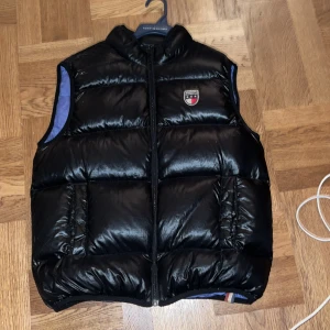 Svart dunväst från Tommy Hilfiger - Säljer en snygg svart dunväst från Tommy Hilfiger. Västen har en dragkedja framtill och ett broderat märke på bröstet. Perfekt för höst och vinter, håller dig varm och stilren. Passar både till vardags och lite finare tillfällen.