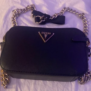 Svart axelväska från Guess - Snygg svart axelväska från Guess med kedjedetaljer och dragkedja. Perfekt för en kväll ute eller en dag på stan. Väskan har en stilren design med Guess-logga framtill.Den är inte använd heller.Den är dessutom äkta.