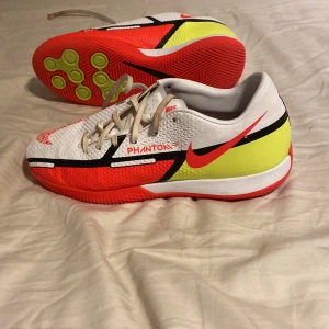 Nike Phantom fotbollsskor - Säljer ett par Nike Phantom fotbollsskor i vitt, rött och gult. Skorna har en snygg design med snörning och är perfekta för fotbollsträning. De är i mycket bra skick och redo för nya äventyr på planen!