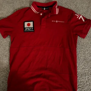 pikétröja från Sail Racing - Snygg röd pikétröja från Sail Racing med vita detaljer och tryck. Den har en klassisk krage med knappar och texten 'JPN77' samt 'Sail Racing International' på både fram- och baksidan. Perfekt för en sportig look! Skick 10/10