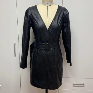 Svart klänning i skinnimitation  - Snygg svart klänning i skinnimitation med v-ringning och bälte i midjan. Den har långa ärmar och en figurnära passform som ger en cool look. Perfekt för en utekväll eller fest! Använt bara en gång som ny.
