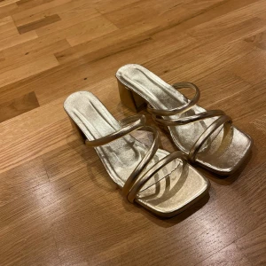 Guldiga sandaler med klack - Säljer ett par snygga guldiga sandaler med klack. De har en fyrkantig tå och smala remmar som korsar över foten. Perfekta för festliga tillfällen och ger en elegant touch till vilken outfit som helst. Endast testade