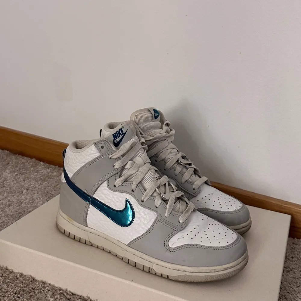 Säljer ett par Nike Dunk sneakers i vitt och grått med en snygg blå swoosh. Skorna har en klassisk design med snörning och är i mycket bra skick. Perfekta för både vardag och festliga tillfällen!. Skor.