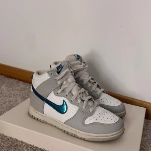 Nike Dunk i vitt och grått med blå detaljer - Säljer ett par Nike Dunk sneakers i vitt och grått med en snygg blå swoosh. Skorna har en klassisk design med snörning och är i mycket bra skick. Perfekta för både vardag och festliga tillfällen!