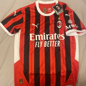 AC Milan fotbollströja från 24/25 säsongen  - Säljer en ny AC Milan fotbollströja i röd och svart med klassiska ränder. Tröjan är kortärmad och har vit krage. Den är i nytt skick med alla etiketter kvar. Perfekt för fans av laget! Stor logga på bröstet och sponsortryck.