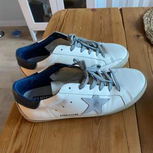 Säljer ett par vita sneakers från Golden Goose i bra skick. De har en grå stjärna på sidan och grå snörning. Skorna har en blå detalj vid hälen och en vit sula. Perfekta för en avslappnad stil!