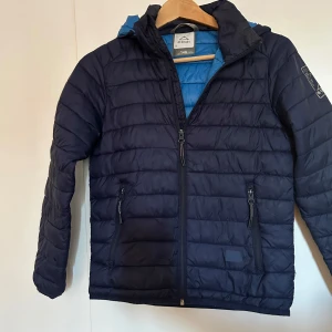 Mörkblå dunjacka från McKinley - Säljer en snygg och varm mörkblå dunjacka från McKinley i storlek 146. Jackan har en dragkedja framtill och två praktiska fickor med dragkedjor. Perfekt för kyliga dagar med sin quiltade design och huva. Passar både till vardags och utomhusaktiviteter.