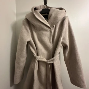 Beige kappa - En superfin beige kappa från New Yorker med en stor huva och knyts med ett bälte i midjan. Perfekt för höst, vinter och vår! Den är skön och varm att ha på sig!! 