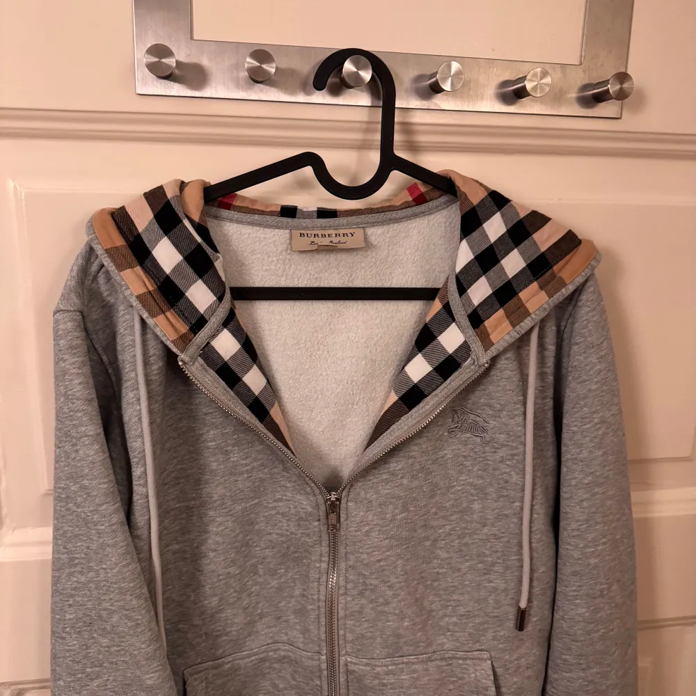 Säljer en snygg grå hoodie från Burberry med klassiskt rutigt mönster i huvan. Den har dragkedja framtill och är i mycket bra skick. Perfekt för höst och vår, och den är supermjuk och bekväm. Passar både till vardags och lite mer uppklädda tillfällen. 🧥✨stl m men passar S. Hoodies.