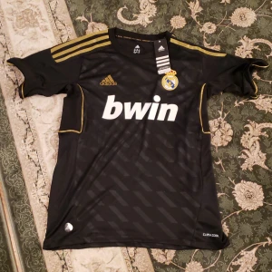 Svart Real Madrid fotbollströja från Adidas - Helt NY! Säljer en svart Real Madrid fotbollströja från Adidas i Climacool-material. Tröjan har korta ärmar med guldiga detaljer och klubbens emblem på bröstet. Perfekt för fans som vill visa sitt stöd med stil! 🖤 priset kan diskuteras