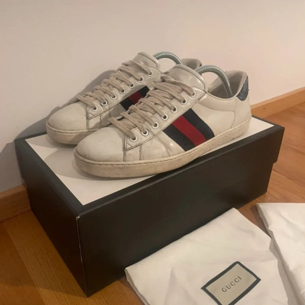 Säljer ett par stiliga Gucci sneakers i vitt skinn med de klassiska ränderna i rött och grönt på sidan. Skorna har snörning och en snygg detalj i svart på hälen. De är i bra skick med lite slitage på sulan. Perfekta för både vardag och fest!. Skor.