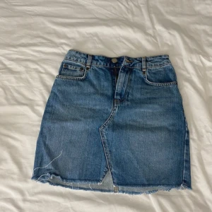 Blå jeanskjol - Säljer en snygg blå jeanskjol i klassisk stil. Kjolen har en rå kant nertill för en trendig look och är perfekt för vardagsbruk. Den har en knapp och dragkedja framtill samt fickor både fram och bak. Passar perfekt till sommaren eller våren!