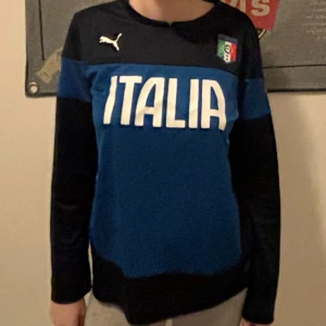 Blå långärmad Italien-tröja från Puma - Säljer en snygg blå långärmad tröja från Puma med texten 'ITALIA' i vitt på framsidan. Tröjan har en mörkare blå nyans upptill och på ärmarna, samt Italiens emblem på bröstet. Perfekt för fotbollsfans! 🏆