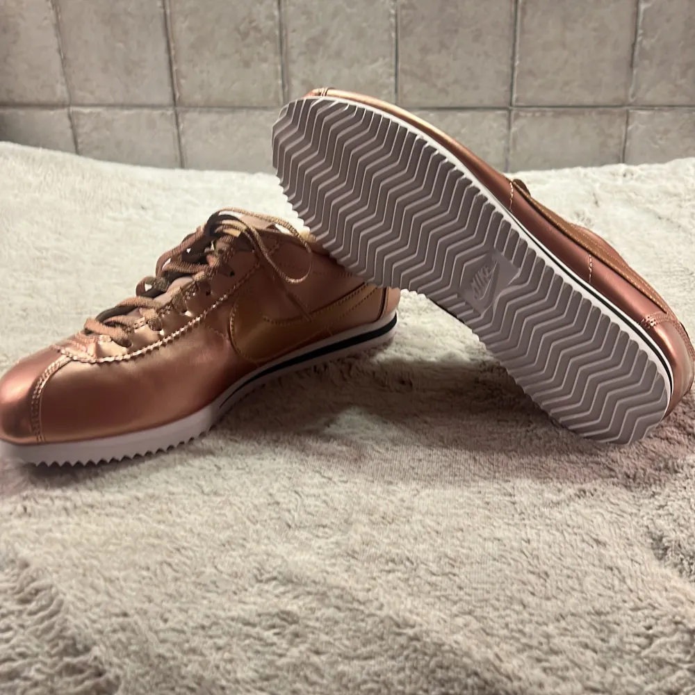 Snygga Nike Cortez sneakers i en glansig roséguld färg. De har en klassisk design med snörning och en vit sula med svart rand. Perfekta för att ge din outfit en extra touch av stil. Skorna är i ny skick och redo för nya äventyr. Storlek 38,5 EU för kvinna. Skor.