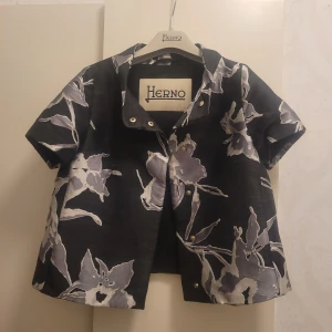 Kortärmad Hernojacka med blommigt mönster - Säljer en superfin och unik blommig jacka från Herno. Den är svart med stora vita och grå blommor, perfekt för att sticka ut lite extra. Jackan har korta ärmar och guldiga knappar framtill. Köpte tyvärr fel storlek så därför jag säljer!🩷 storlek 38 IT som motsvarar XS men sitter litet på mig som vanligtvis har XS och S så passar nog mer XXS och XS🩷 en skyddande påse från Herno följer med och kan skicka med galgen till extra betalning då den hör till en annan Herno jacka jag har!!🥰 jättebra skick