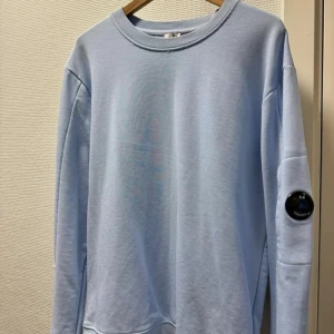 C.P COMPANY sweater - Tidlös klassisk sweater från italienska C.P COMPANY, med den ikoniska linsen och fickan på vänster arm. Inköpt på NK herrtrend 2023 för 2199:-. Mycket sparsamt använd. Tröjans enda defekt är märkeslappen (som självklart inte syns vid användning) i nacken, se bild.  Intresserad av snabb och seriös affär.