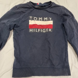 Mörkblå tröja från Tommy Hilfiger - Säljer en snygg mörkblå tröja från Tommy Hilfiger med deras ikoniska logga i vitt och rött på framsidan. Tröjan har långa ärmar och ribbade muddar vid ärmslut och nederkant. Perfekt för en casual look!