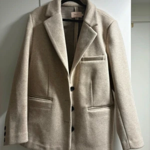 Beige kappa från Nelly - Säljer en stilren beige kappa från Nelly i mycket bra skick. Den har en klassisk design med knappar framtill och fickor på sidorna. Perfekt för höst och vår, och ger en elegant touch till vilken outfit som helst.