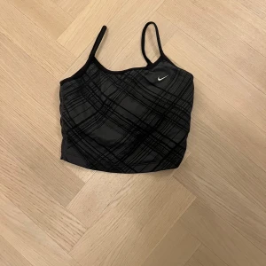 Svart och grå linnetopp från Nike - Snygg och sportig svart och grå linnetopp från Nike med ett rutigt mönster. Perfekt för träning eller en avslappnad dag. Toppen har smala axelband och är i mycket bra skick. Passar perfekt till både gymmet och vardagsoutfiten! Den är i storlek M men passar även S