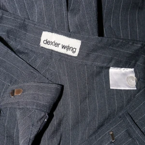 Randiga kostymbyxor från Dexter Wong - Snygga och stilrena kostymbyxor från Dexter Wong i mörkgrått med tunna vita ränder. Byxorna har en klassisk design med hög midja och vida ben, perfekt för både kontoret och festligare tillfällen. Materialet känns lyxigt och bekvämt. Ett måste för garderoben!