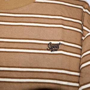 Brun och vit randig tröja från Sweet SKTBS - Säljer en snygg brun och vit randig tröja från Sweet SKTBS. Den har en avslappnad passform och korta ärmar, perfekt för en casual look. Tröjan är i bra skick och har en broderad logga på bröstet. Perfekt för vår och sommar!