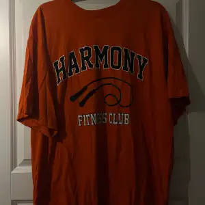 Säljer en orange t-shirt med texten 'Harmony Fitness Club' tryckt på framsidan. Perfekt för träning eller en avslappnad dag. T-shirten är i mycket bra skick och har en oversize passform.