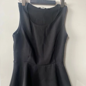 Svart ärmlös klänning - Säljer en elegant svart topp med en stilren design. Den är ärmlös och har en figurnära passform upptill och utsvängd volang nertill. Perfekt för festliga tillfällen eller en utekväll. Materialet känns mjukt och bekvämt mot huden.