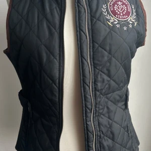 Svart quiltad väst från U.S. Polo Assn. - Säljer en snygg svart quiltad väst från U.S. Polo Assn. med ett broderat emblem på bröstet. Västen har en dragkedja framtill och knappar i sidan för justering. Perfekt för höst och vår!