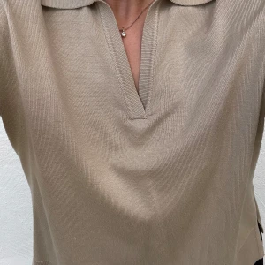 Beige pikétröja - Säljer en stilren beige tröja i normal passform. Tröjan har korta ärmar och en klassisk krage med en liten v-ringning, slits på båda sidor. Perfekt för både vardag och lite mer uppklädda tillfällen. Superbekväm och lätt att matcha med olika outfits!