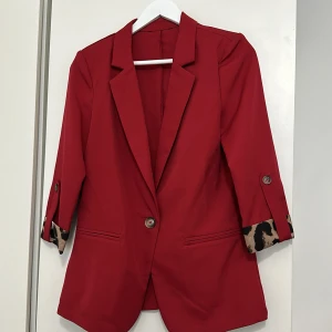 Röd kavaj med leoparddetaljer - Säljer en snygg röd kavaj med leopardmönstrade detaljer på ärmsluten. Kavajen har långa ärmar och knäpps med knappar framtill. Perfekt för att ge en färgklick till din outfit!