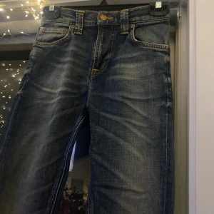 Jeans från Nudie Jeans - Säljer ett par snygga jeans från Nudie Jeans. De är i en klassisk blå färg med snygga slitningar och kontrastsömmar i orange. Perfekta för en casual look och passar till det mesta. De har en knappgylf och fem fickor. Det är skinny jeans 