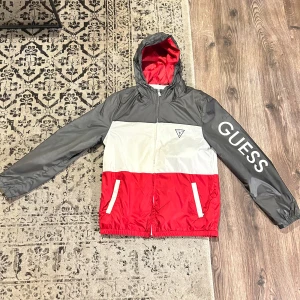 Flerfärgad vindjacka från Guess - Säljer en snygg och stilren vindjacka från Guess i storlek M. Jackan har en cool färgblockdesign i svart, vit och röd med Guess-loggan tryckt på ärmen. Den har en dragkedja framtill och praktiska fickor. Perfekt för vår och höst!
