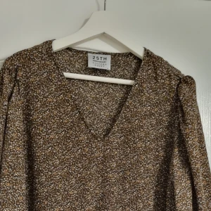 Brun mönstrad klänning från 25TH - Säljer en superfin brun klänning från 25TH med ett diskret mönster. Den har en v-ringad hals och långa ärmar, perfekt för höst och vinter. Klänningen är i ett mjukt och bekvämt material, vilket gör den idealisk för både vardag och fest.