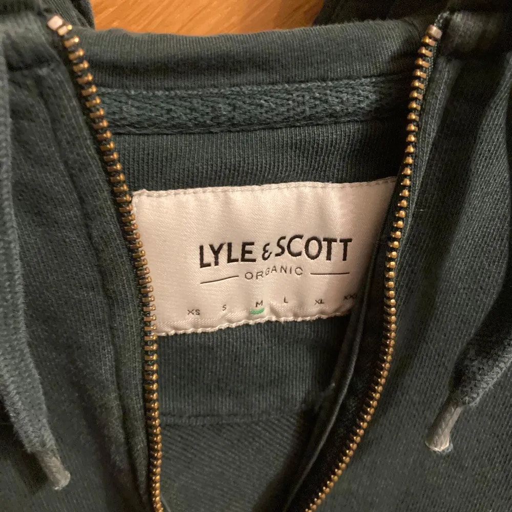 Säljer en snygg grön hoodie från Lyle & Scott i storlek M. Den är gjord av ekologiskt material och har en dragkedja framtill. Perfekt för en avslappnad stil med sin klassiska design och det ikoniska märket på bröstet. Passar både till vardags och chillkvällar. 🐥. Tröjor & Koftor.