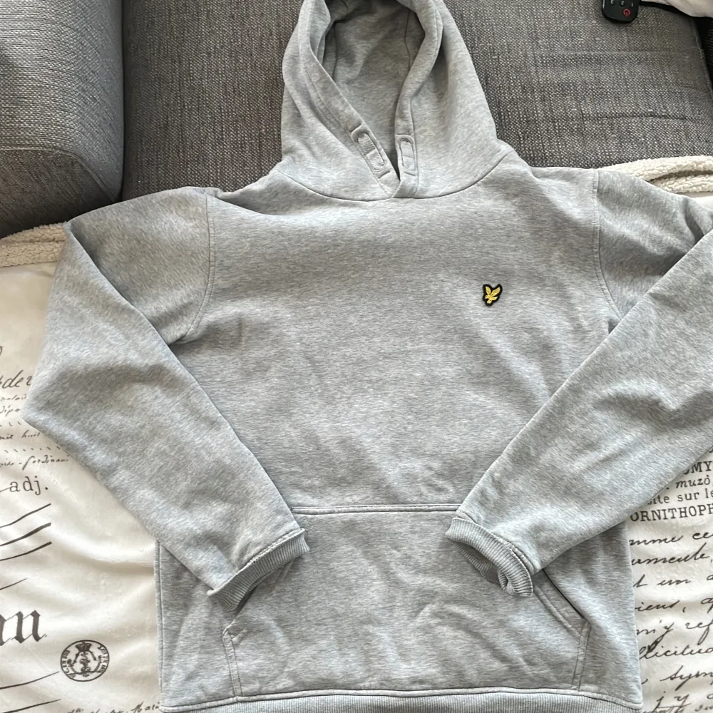 Säljer en grå hoodie frå Lyle& Scott Hoodien har en klassisk passform med ribbade muddar vid ärmslut och nederkant.  Ärm muddarna undersida har förslitning se bild men syns inte påDen har en stor ficka framtill och en bekväm huva. . Tröjor & Koftor.