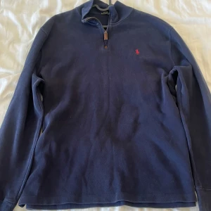 Mörkblå tröja från Polo Ralph Lauren - Säljer en snygg mörkblå halfzip från Polo Ralph Lauren i storlek XL. Tröjan har en halv dragkedja och det klassiska röda Polo-loggan broderad på bröstet. Perfekt för höst och vinter med sin stilrena design och bekväma passform. Passar både till vardags och lite finare tillfällen.