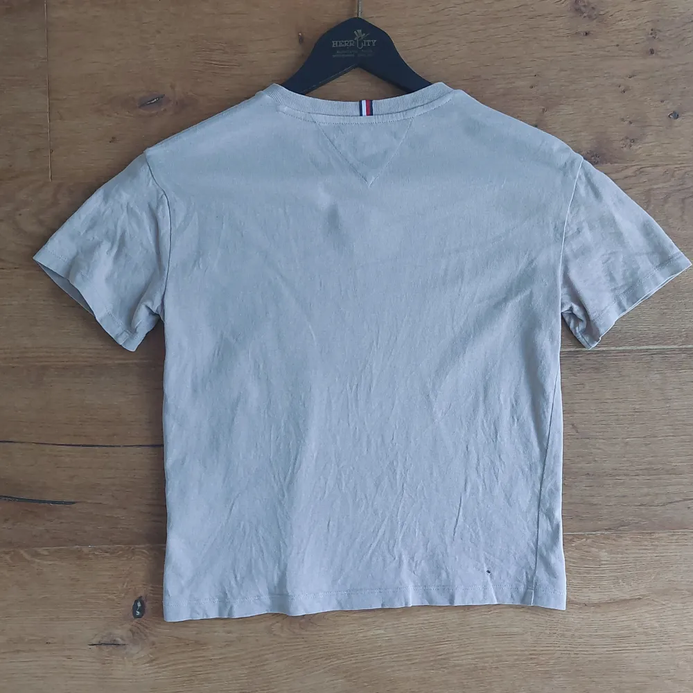 Säljer en snygg beige t-shirt från Tommy Hilfiger. Den har en klassisk rund hals och korta ärmar. På framsidan finns en broderad Tommy-logga i rött, vitt och blått. Perfekt för en avslappnad stil! OPS har två små hål på framsida  och ett på baksidan.. T-shirts.