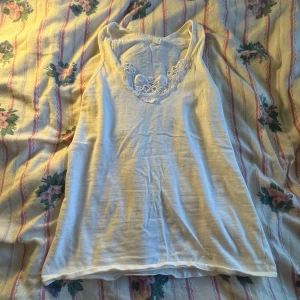 Vit linne/topp - Säljer en söt vit ärmlös topp med vacker spetsdetalj vid halsringningen. Perfekt för sommardagar eller som ett lager under en kofta. Toppen är i ett mjukt och bekvämt material.