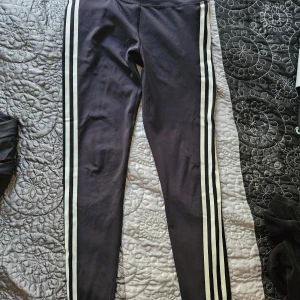 Svarta leggings från Adidas - Säljer ett par svarta leggings från Adidas med vita ränder längs sidorna. De är i storlek M och passar perfekt för träning eller en avslappnad dag. Materialet är stretchigt och bekvämt.