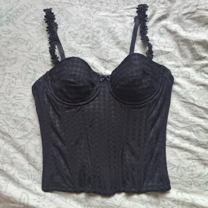 Svart korsett top - Svart korsett topp med blommor på axelbanden. Rutigt mönster med en rosett detalj. Thriftat men från märket Marie Jo. Storlek 70B