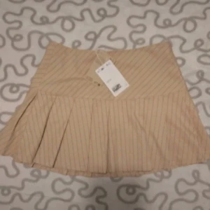 Beige randig kjol från Monki - Säljer en superfin beige kjol från Monki i storlek 38. Den har ett snyggt randigt mönster och är perfekt för både vardag och fest. Kjolen är kort och har en härlig passform som ger en chic look. Den är i nyskick och har fortfarande prislappen kvar!