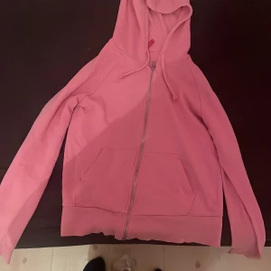 Rosa hoodie med dragkedja - Säljer en mysig rosa hoodie i storlek 38. Den har en dragkedja framtill och en praktisk ficka. Perfekt för en avslappnad stil eller kyliga dagar. Huvan ger extra värme och komfort.